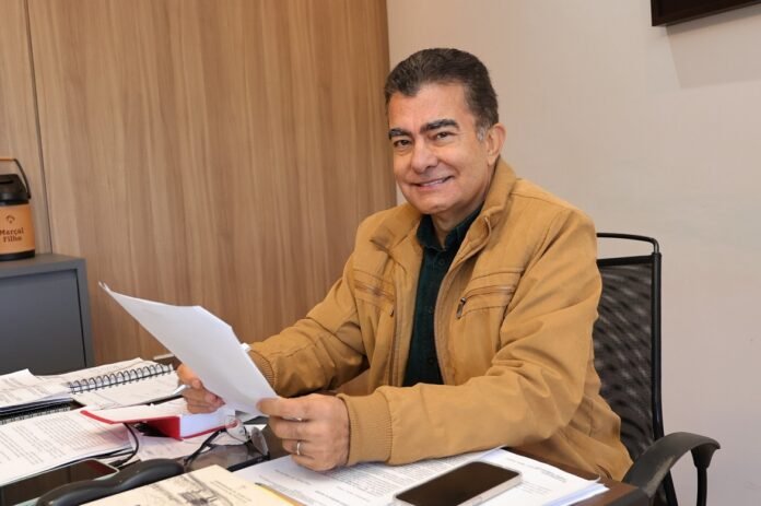 Man in a tan jacket sits at a cluttered office desk, smiling while reviewing a sheet of paper.