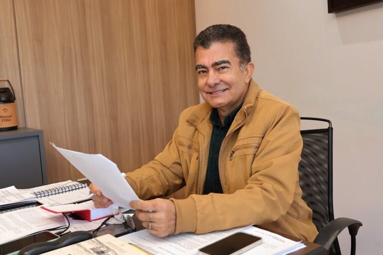 Man in a tan jacket sits at a cluttered office desk, smiling while reviewing a sheet of paper.