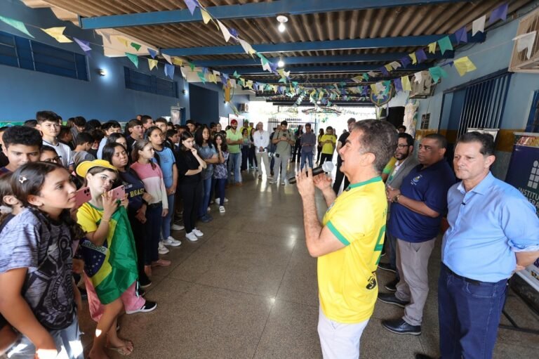 Man in a yellow shirt speaks into a microphone to a large group of students gathered in a festively decorated hallway with colorful bunting overhead.