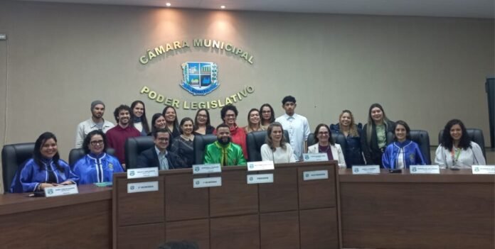 Group of people in a city council chamber, sitting and standing behind a long desk with nameplates and the municipal seal on the wall behind them is visible.