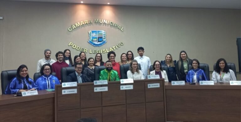 Group of people in a city council chamber, sitting and standing behind a long desk with nameplates and the municipal seal on the wall behind them is visible.",