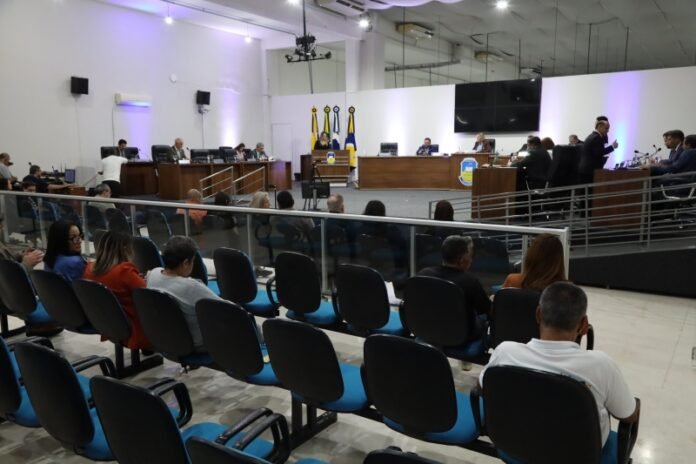 Government council chamber: officials seated at a long dais with flags behind, audience in rows watching a formal meeting