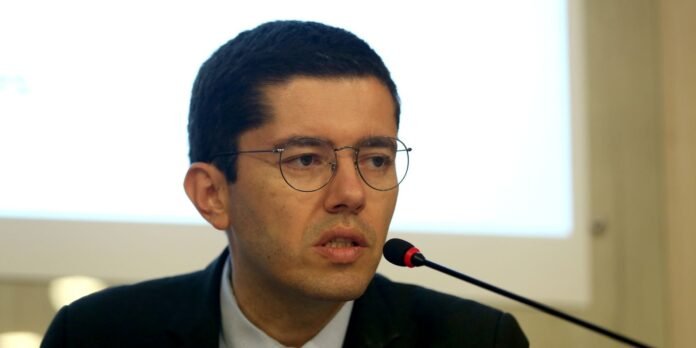 Man in a suit and round glasses speaks into a microphone at a conference conference panel](#)