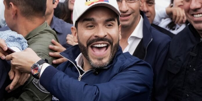 Smiling man in a navy jacket and white cap in a cheering crowd of people around him.