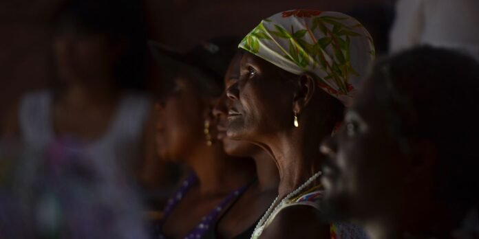 Profile of a dark-skinned woman in a white headscarf with green leaf pattern, gold earring, and pearl necklace, seated among a dimly lit crowd