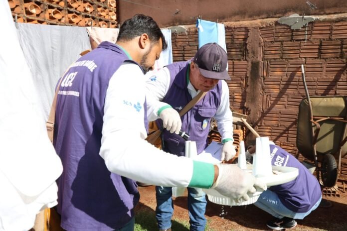Two field workers in matching purple vests and gloves assemble a white plastic filtration device outdoors beside a brick wall.