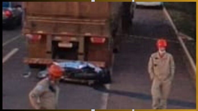 Two workers in orange hard hats stand near a damaged blue car beneath a large brown truck on a roadway.