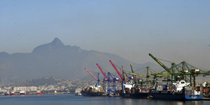 Port scene with colorful container cranes and stacked shipping containers docked along the quay, with a mountain range in the hazy background.
