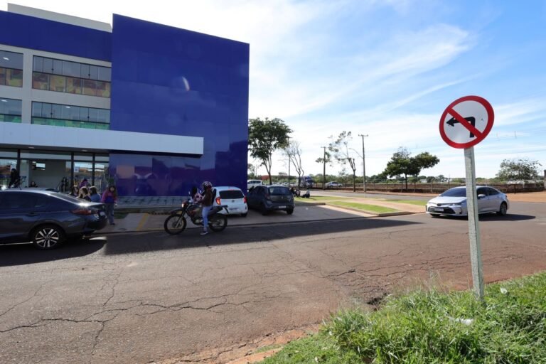 Front view of a blue/purple building with people near the entrance, several parked cars, and a motorcyclist waiting by a bike; a 'no left turn' sign is on the right side of the image.