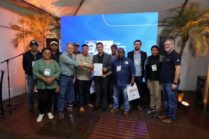 Group of people on a stage posing with a certificate in front of a blue display screen and palm trees in the background.