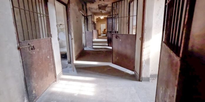 Dusty prison corridor with open cell doors and barred walls, sunlight streaming through.