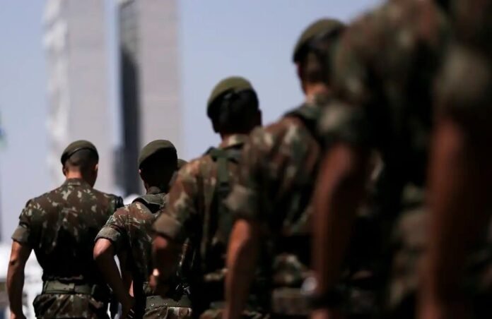 Line of soldiers in camouflage uniforms standing in formation outdoors, backs to the camera, helmets on.