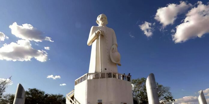 Statue of a tall white man holding a cane and hat atop a pedestal under a blue sky with clouds, overlooking a viewing platform.