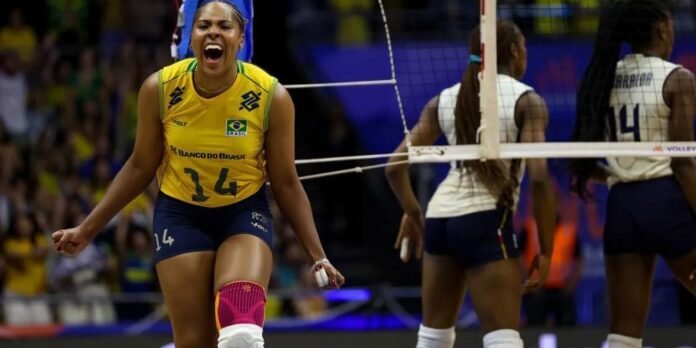 Brazilian volleyball player in yellow jersey number 14 celebrates on the court with a pink knee pad and clenched fist, net in the background.