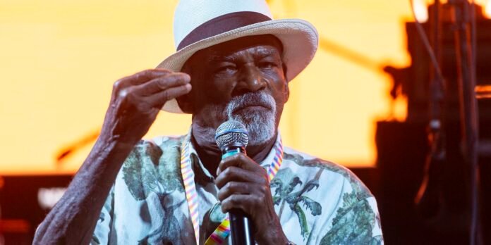 Older man with a white hat and beard speaks into a handheld microphone on a brightly lit stage wearing a tropical shirt.
