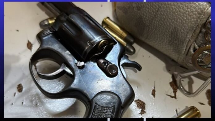 Close-up of a revolver on a table with spent bullet casings nearby and a gray purse in the background.