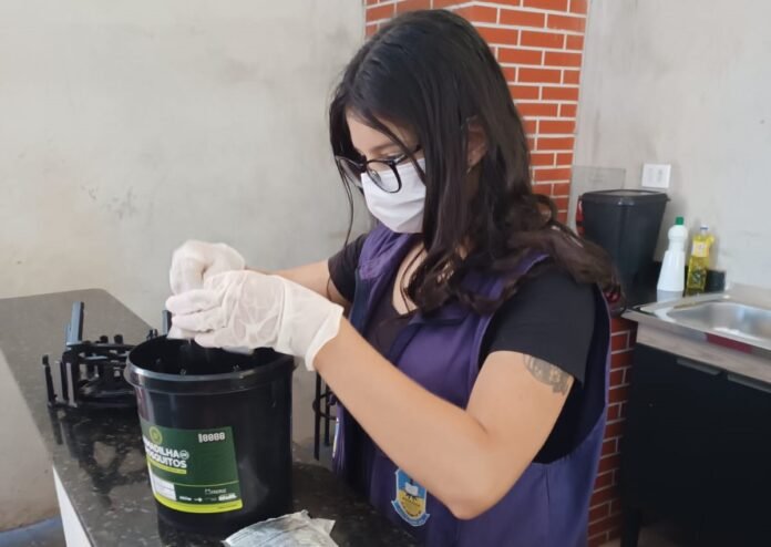Woman wearing a face mask and white gloves handling the contents of a black plastic bucket on a kitchen counter, with a stove nearby and a brick wall backdrop.