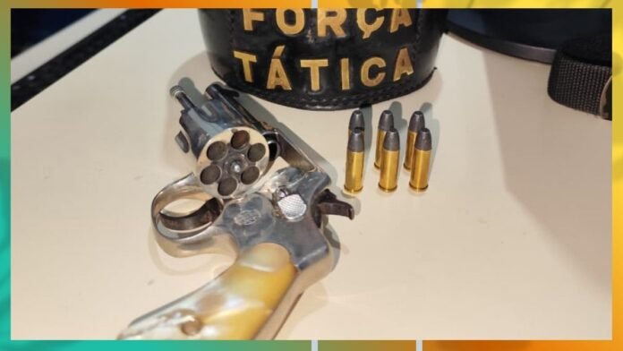 A revolver with a six-shot cylinder on a table, three bullets standing upright, and a tactical helmet visible in the background reading ‘FORÇA TÁTICA’.