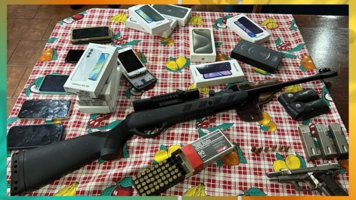 Assortment of boxed smartphones and devices spread on a checkered tablecloth beside a black rifle and ammunition on the table.