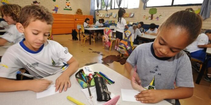 Two young students sit at a round table drawing on white paper, with markers and a pencil case in the middle, in a colorful classroom.