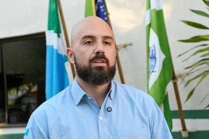 Bearded man in a light blue button-down shirt stands outdoors, with green and blue flags in the background.