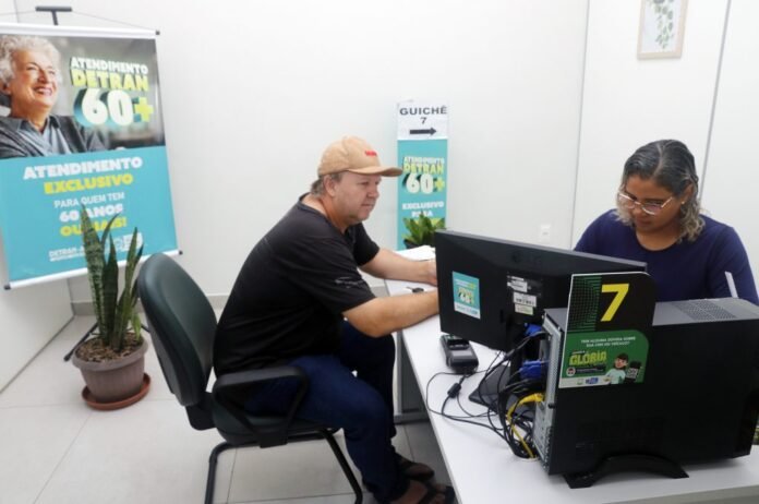 Two people sit at a service desk with a computer; posters advertise 60+ atendimento in a waiting area in the background.