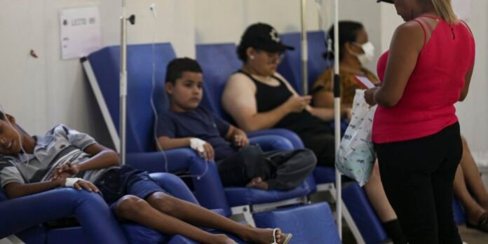 People in a clinic donating blood on blue reclining chairs, with IV poles nearby; a woman in a pink top stands holding a bag and papers in the foreground.