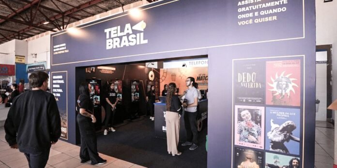 Exhibition booth with a large blue wall reading 'TELA BRASIL' and people talking near a service counter inside a dark display hall.