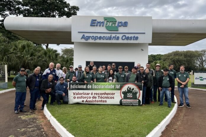 Group of people posing outdoors in front of the Embrapa Agropecuária Oeste sign, holding a banner advocating higher education for technicians and assistants.