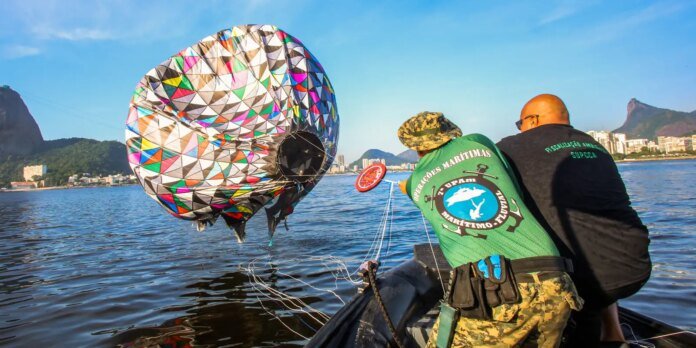 Two men in a small boat deploy a colorful parachute-like device into the water near a coastal city.