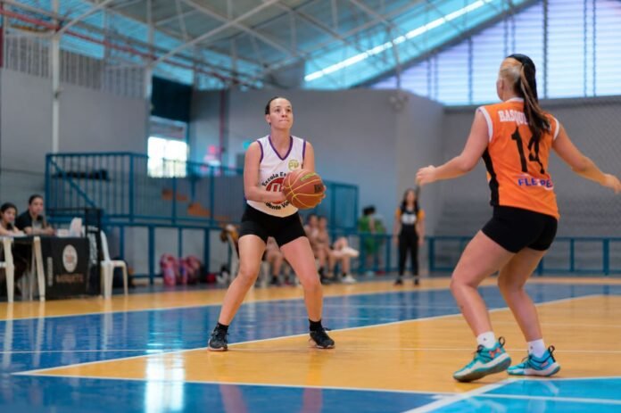 Female basketball player in a white jersey holds the ball while being guarded by an opponent in orange on a bright indoor court with spectators in the background.