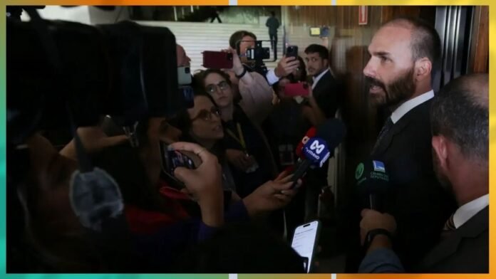 Bearded man in a suit is interviewed by multiple reporters holding microphones and smartphones in a crowded press area.