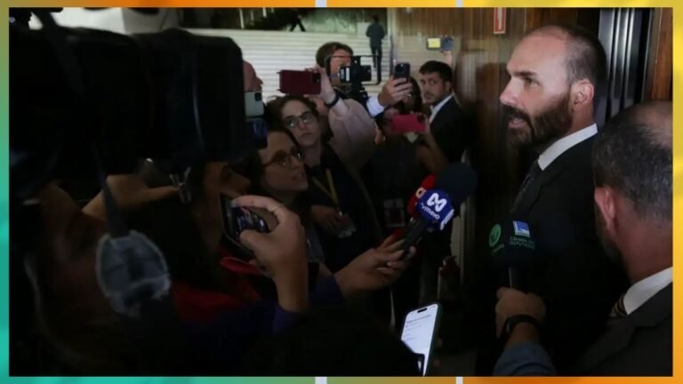 Bearded man in a suit is interviewed by multiple reporters holding microphones and smartphones in a crowded press area.