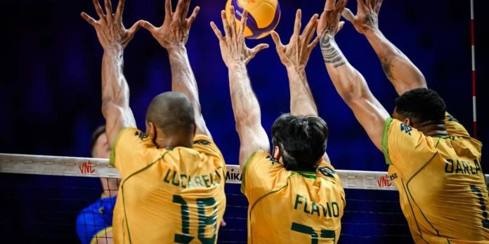 Four volleyball players in yellow jerseys jump with arms raised at the net to block a ball during a match at the net edge.