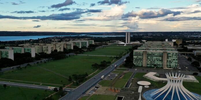 Aerial view of a modern campus with green glass buildings, a large central lawn, roads, and a white sculptural pavilion by a pool, with a lake in the background.