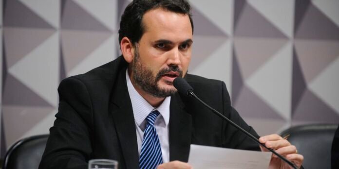 Man in a dark suit and striped tie speaking into a microphone at a panel, holding a sheet of paper
