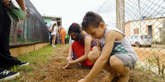 Two kids crouch in a dirt patch beside a chain-link fence, planting seeds with their fingers as adults work nearby in a garden setting.