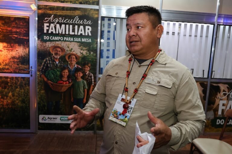 Man in a beige utility shirt and beaded necklace speaks or gesturing at an event, with a large poster behind him reading 'Agricultura Familiar' and showing a farmer family with produce in a basket.