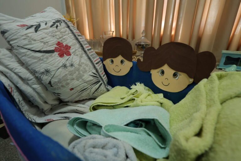 A child's bed with a floral quilted pillow, pastel blankets, and two smiling cartoon-face cutouts leaning against the pillows.