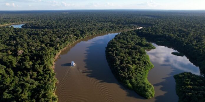 Aerial view of a winding muddy river bordered by dense green forest, with a small boat creating ripples in the water.