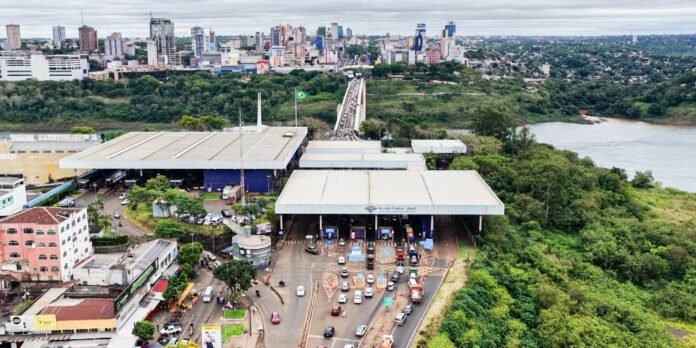 Aerial view of a river, a busy toll plaza with cars, and a city skyline in the distance amid green riverbanks.