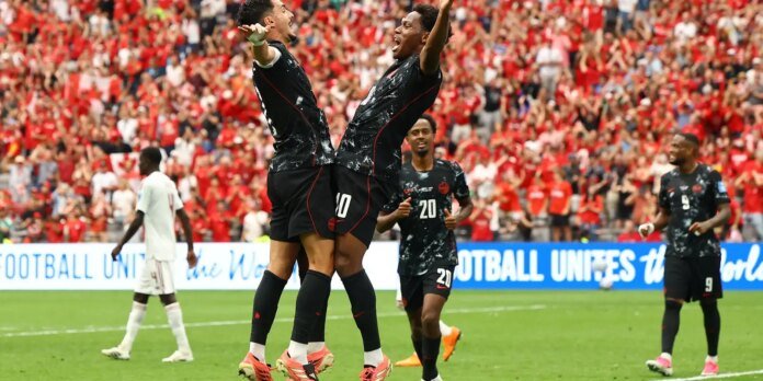 Soccer players in black uniforms celebrate a goal, jumping and embracing on the field as a red-clad crowd cheers in the stands behind them.