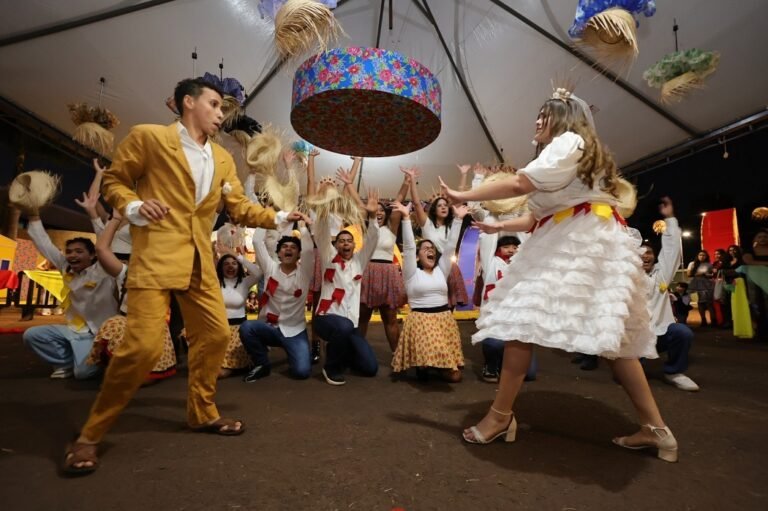 People dancing at a festive party, a man in a yellow suit and a woman in a white frilly dress lead the crowd under a colorful hanging banner.