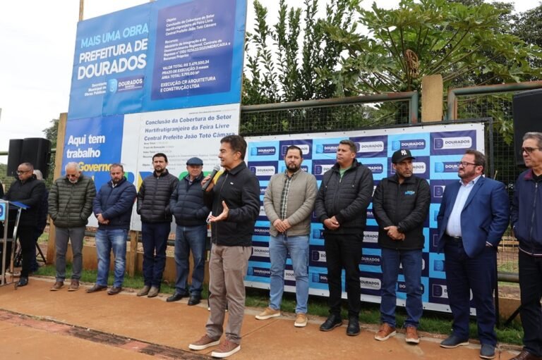Group of men lined up outdoors at a press event, one man speaks into a microphone, blue banners behind them read Dourados/Prefeitura de Dourados.
