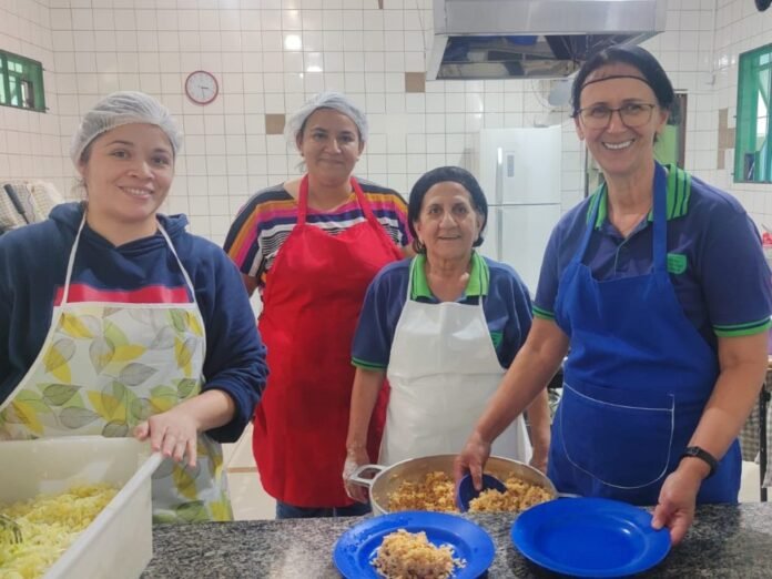 Four women in a bright kitchen wearing aprons and hairnets, smiling as they prepare and plate a dish together.