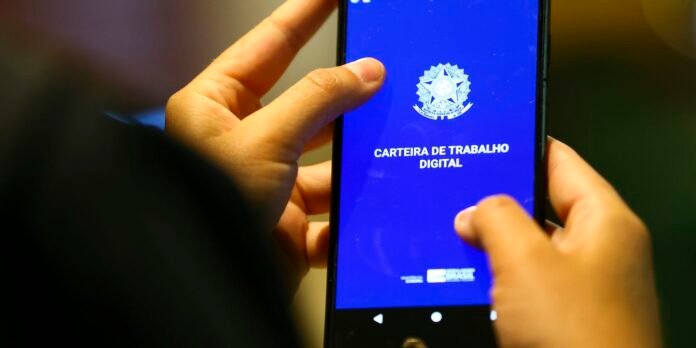 Close-up of hands holding a smartphone displaying the Brazilian Carteira de Trabalho Digital app on a blue screen with an official seal above the text, indicating the digital work card.