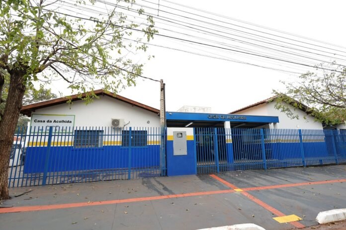 Blue metal fence and gate enclose a white, single-story building with blue and yellow trim labeled 'Casa da Acolhida' (shelter) along a street with trees and overhead power lines behind it
