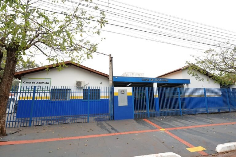 Blue metal fence and gate enclose a white, single-story building with blue and yellow trim labeled 'Casa da Acolhida' (shelter) along a street with trees and overhead power lines behind it