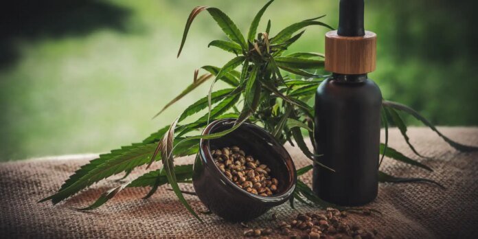 Cannabis leaves with a jar of hemp seeds and a dropper bottle on burlap outdoors.
