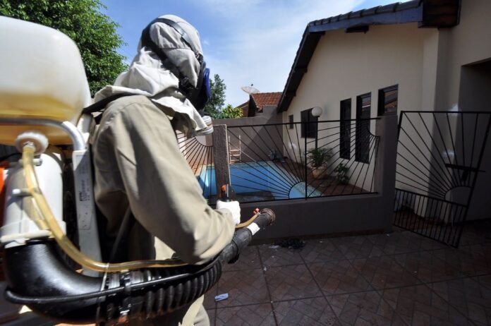 Worker in full protective hazmat suit with backpack sprayer spraying near a house and pool area in a gated patio.